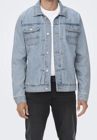 Ljusblå denimjacka med knapstängning, två bröstfickor och en avslappnad design. Bärs över en vit T-shirt och svarta jeans.