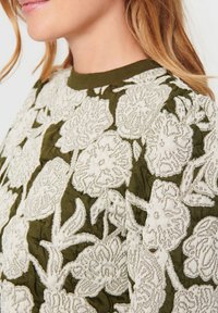 Grüner Stoff mit filigraner weißer Blumenstickerei, strukturierten Details, geripptem Halsauschnitt und sichtbaren Nähakzenten.