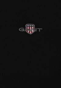 Black cotton fabric featuring an embroidered logo with a shield design in grey, red, and blue, displaying the word "GANT."