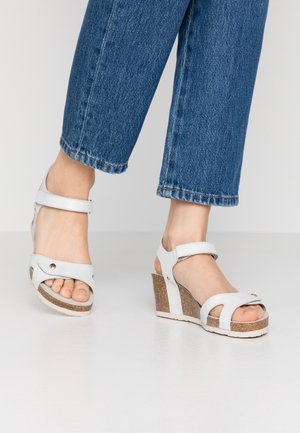 Sandales blanches à talons compensés avec une bride à la cheville, semelle en liège, design ouvert à l'avant et accents en métal argenté. Associées à un pantalon cropped en denim bleu.