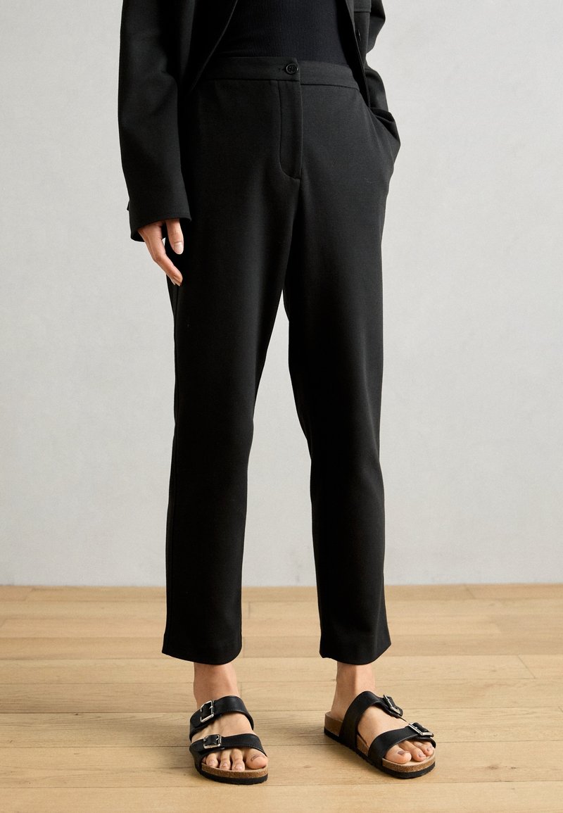 Personne portant un pantalon noir sur mesure et des sandales noires à boucle, debout sur un sol en bois devant un mur uni.