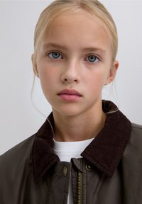 Giacca verde scuro con colletto in velluto a coste marrone, chiusura con cerniera e accessori dorati, indossata sopra una camicia bianca. Tessuto dalla texture liscia.