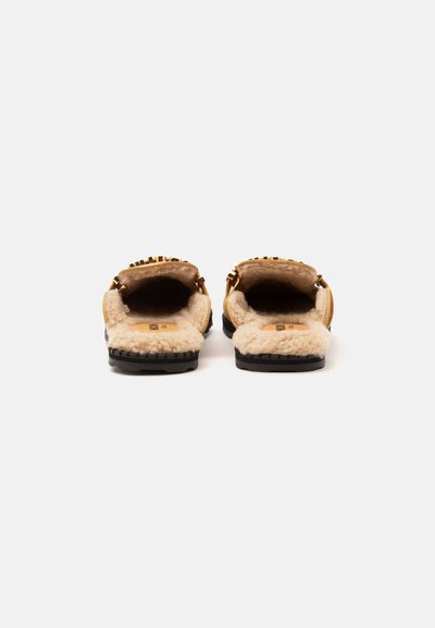 Paire de chaussons marron clair à enfiler avec doublure intérieure crème duveteuse et semelles noires texturées vues de dos sur fond blanc.