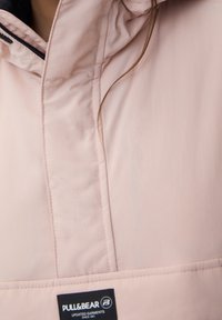 Helle rosa Jacke mit Reißverschluss und hohem Kragen, mit einem schwarzen Innenfutter und einem Etikett am unteren Rand mit "PULL&BEAR" Branding.