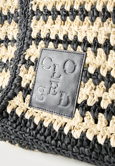 Υφαντό ύφασμα σε μαύρο και μπεζ με υφή πλεκτού, με ένα ραμμένο τετράγωνο έμπλαστρο από μαύρο δέρμα, με ανάγλυφα γράμματα που σχηματίζουν τη λέξη "ΚΛΕΙΣΤΟ" σε μοτίβο σταυρού.