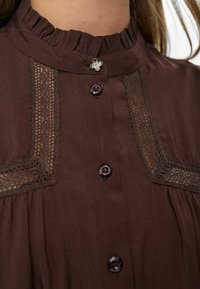 Bruine blouse met kantdetails, gerimpelde kraag, donkere knopen en een kleine bloemetjespin nabij de halslijn.