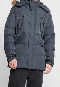 Chaqueta acolchada gris con capucha adornada con pelo, bolsillos en el pecho con cremallera y cierres de presión. Cuenta con puños elásticos y un patrón acolchado.