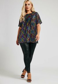 Top con lentejuelas de purpurina multicolor sobre tela negra, de mangas cortas y corte suelto, combinado con leggings de cuero sintético negro y sandalias de tacón.