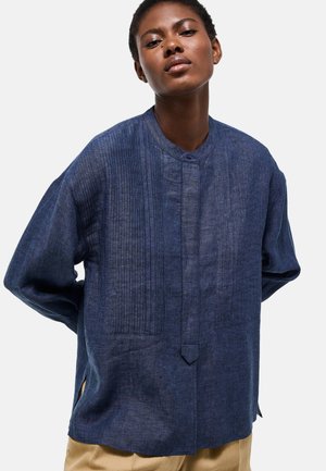 Femme portant une chemise ample bleu marine texturée à manches longues avec des plis verticaux et un pantalon beige, debout les mains dans le dos.