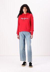 Sarkana kapuce ar "Pepe Jeans" logotipu, brīva piegriezuma, ar auklu, ribotām piedurknēm, saskaņota ar gaiši ziliem taisniem džinsiem un melniem sporta apaviem.