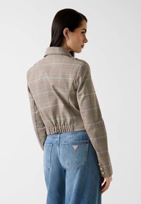 Veste courte à carreaux marron avec col, taille élastique et boutons dorés en accent, associée à un jean taille haute bleu clair.
