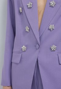 Blazer lilla con colletto a revers, due tasche frontali e sei decorazioni in cristallo disposte simmetricamente sul davanti.