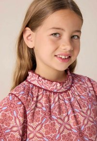 Rosafarbene Bluse mit rüschigem Ausschnitt, die florale und geometrische Muster in Rot, Blau und Gelb aufweist. Glatte Stofftextur.