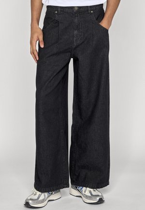 Personne portant un jean en denim noir à jambes larges et des baskets blanches avec des accents bleus et gris, debout devant un fond clair uni.