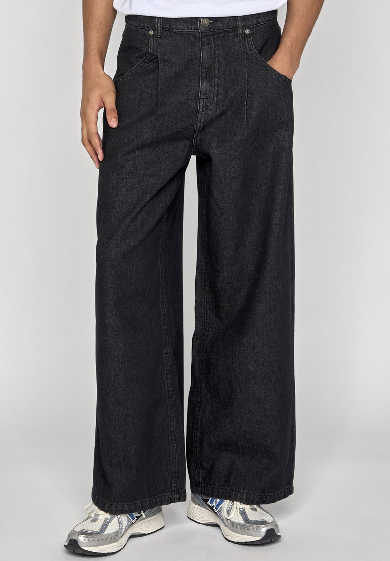 Personne portant un jean en denim noir à jambes larges et des baskets blanches avec des accents bleus et gris, debout devant un fond clair uni.