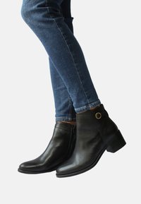 Schwarze Leder-Ankle-Boots mit einem niedrigen Absatz, ausgestattet mit einem seitlichen Reißverschluss und einem goldenen Akzent am Knöchel. Getragen mit dunkelblauen Skinny Jeans.