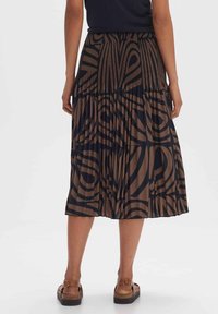 Kvinna som bär en brun och svart abstrakt mönstrad plisserad midi-kjol med bruna spännskor, stående mot en enfärgad bakgrund.