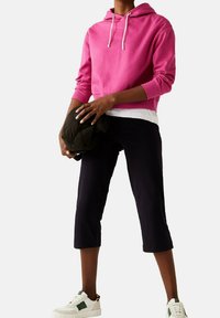 Sudadera con capucha rosa con cordones, camisa blanca de varias capas debajo, pantalones oscuros recortados y zapatillas blancas con detalles en verde. Textura suave y estilo casual.