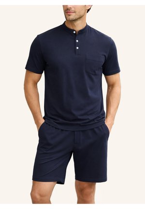 Homme portant un tee-shirt henley bleu marine à manches courtes avec trois boutons et une poche poitrine, associé à un short bleu marine assorti, mains dans les poches.