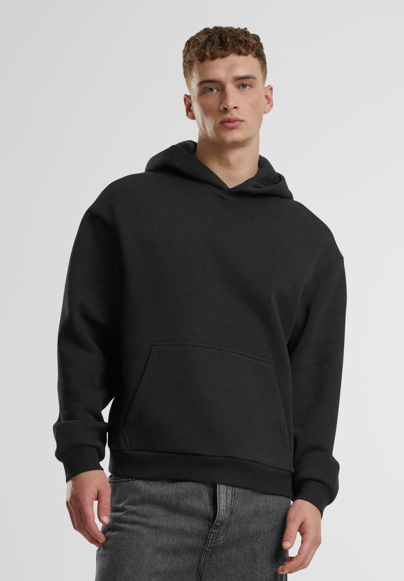 Junger Mann mit lockigem Haar, der einen schlichten schwarzen Hoodie und graue Jeans trägt und vor einem hellgrauen Hintergrund steht.