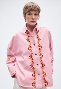 Ροζ φαρδιά πουκάμισο με μαύρα κουμπιά, διακοσμημένο με ζωντανή πορτοκαλί και κόκκινη γεωμετρική κεντητική μπροστά και στα μανίκια. Μαλακό υφιστό με υφή.