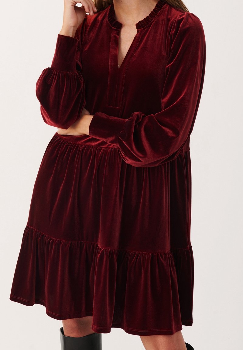 Femme portant une robe en velours bordeaux à manches longues, à volants, avec un col en V et des poignets froncés, associée à des bottes noires montantes jusqu'aux genoux.