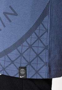 Blue athletic shirt with a textured finish, featuring geometric black patterns and a branded tag with Paris Saint-Germain and Nike logos.