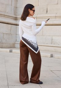 Bela rebrasta pulover s off-shoulder dizajnom, ki ima velik črno-beli poudarek s pikami. Rjave široke hlače, gladke teksture.