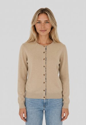 Blonde vrouw met een beige vest met knopen over een top met patroon en lichtblauwe spijkerbroek, staand tegen een effen lichte achtergrond.