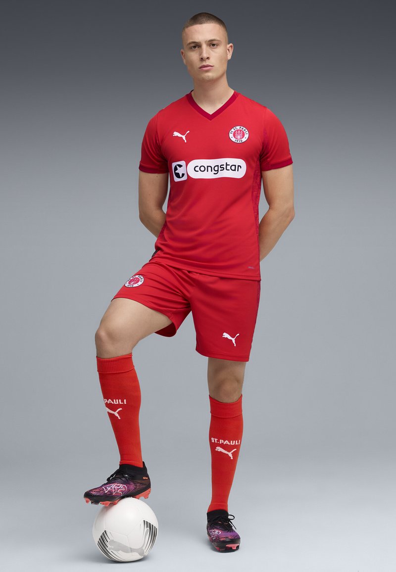 Röd fotbollsklänning med korta ärmar och shorts, med en v-ringning, logotypdetaljer och matchande röda strumpor. Svarta och lila fotbollsskor.