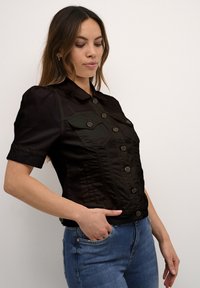 Svart kortärmad jacka i glänsande tyg, med krage, knäppning fram och två bröstfickor. Visar en avslappnad passform.