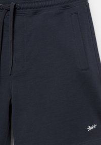 Gros plan d’un pantalon de jogging bleu foncé avec taille à cordon, poches latérales et petit logo blanc brodé près de l’ourlet.