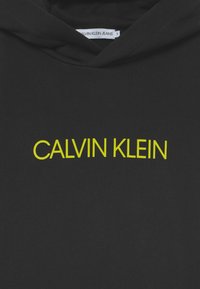 Felpa nera con cappuccio caratterizzata da un logo giallo "CALVIN KLEIN" in un carattere audace. Texture morbida, adatta per un abbigliamento casual.