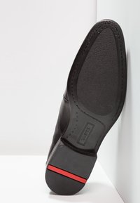 Chaussure en cuir noir avec un dessus lisse, une semelle texturée et une touche de rouge sur le talon. Présente un bout rond et une couture détaillée.
