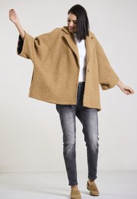 Tan, textured, oversized coat with a collar, two buttons, and wide sleeves; paired with dark jeans and tan shoes.