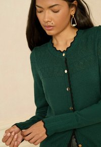 Zelený kardigán s textúrovaným pleteným dizajnom, ryhovanými okrajmi, čiernymi akcentmi a zlatými gombíkmi. Dlhé rukávy s priliehavými manžetami.