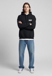 Felpa nera con testo "AMOUR", in misto cotone, con tasche frontali. Abbinata a jeans azzurri e scarpe da ginnastica bianche. Modello in piedi con le braccia incrociate.