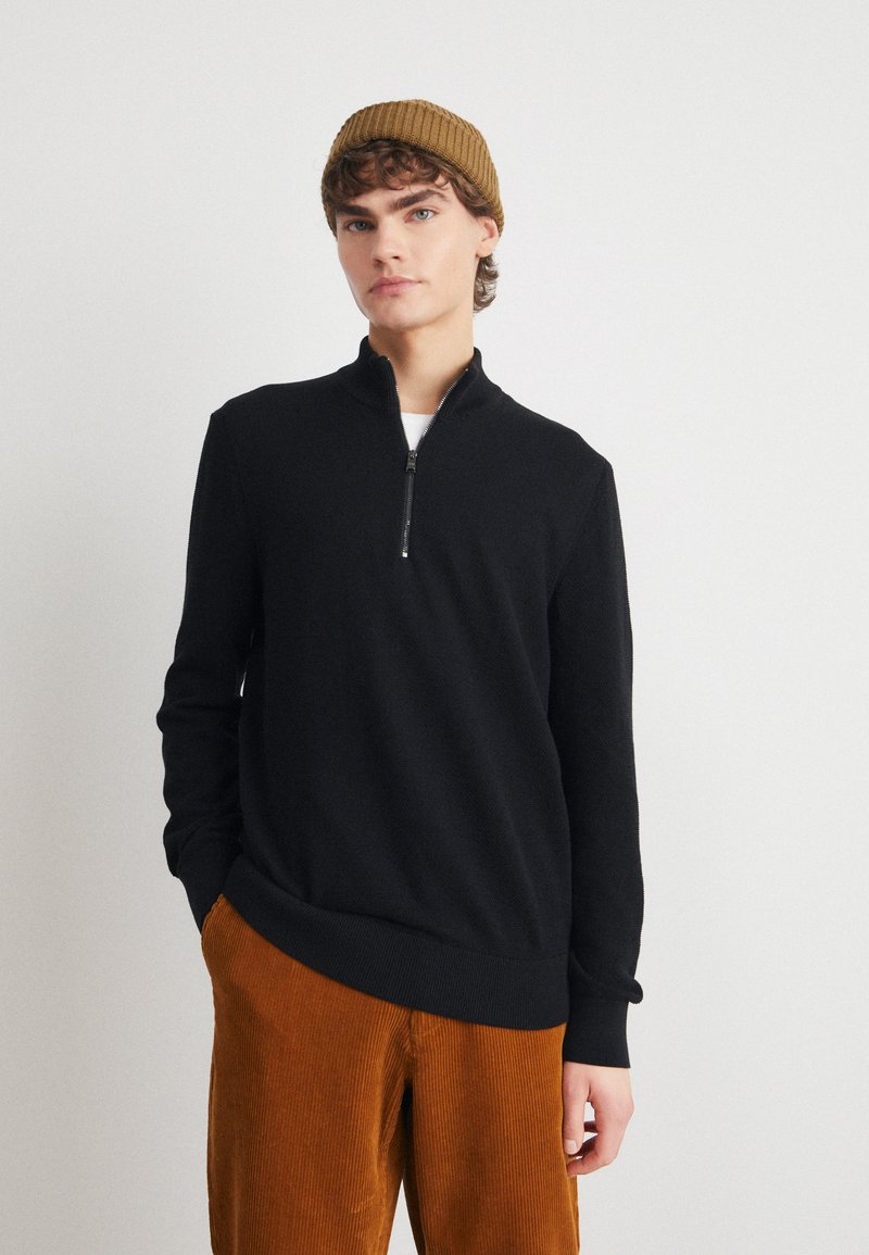 Black zip-neck sweater in soft fabric, featuring long sleeves and a ribbed hem. Paired with brown corduroy pants.