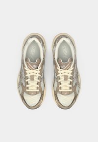 Zapatillas deportivas en tonos beige y gris con materiales de malla y cuero, que presentan una puntera redondeada, lengüeta acolchada, cordones beige y detalles con logotipo.