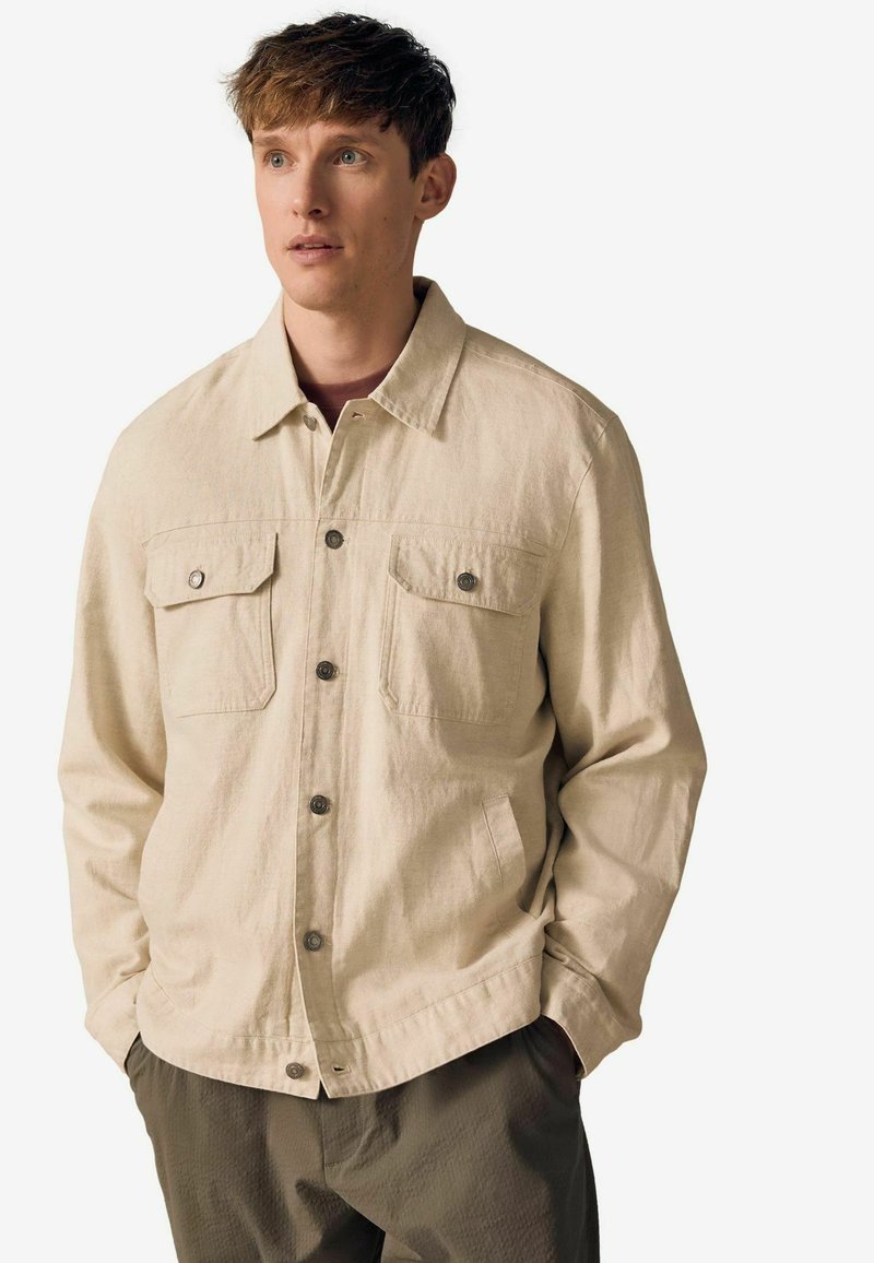 Veste boutonnée beige en tissu texturé. Présente deux poches poitrine avec rabats et une coupe décontractée. Portée par-dessus un t-shirt noir à col rond.