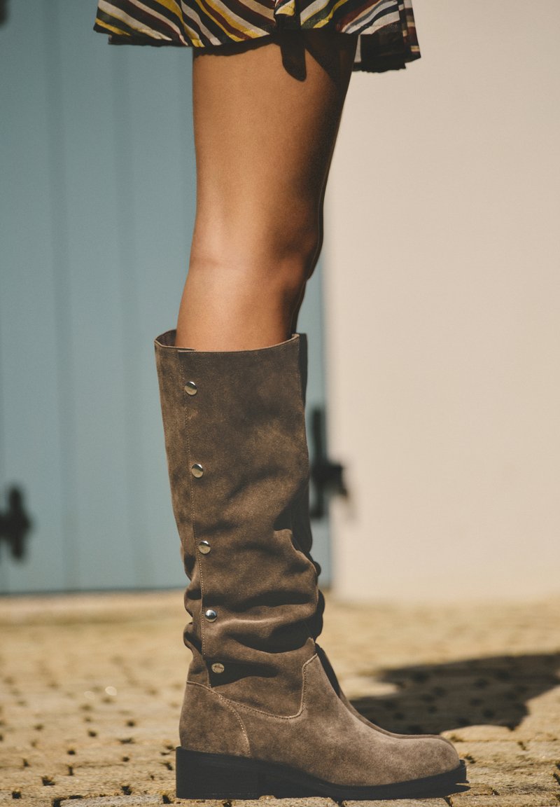 Une jambe portant des bottes hautes en daim marron avec des boutons métalliques, associée à une jupe rayée, debout sur un pavé en pierre.