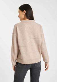 Beige gebreide trui met een kraaghalslijn, losse pasvorm, geribbelde zoom en subtiele horizontale stikdetails. Gecombineerd met donkere jeans.