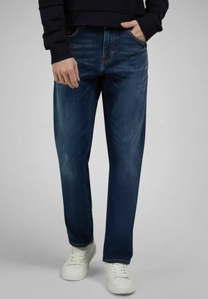 Osoba ubrana w ciemnoniebieskie dżinsy, białe trampki i czarną bluzę z długim rękawem, stojąca z jedną ręką w kieszeni na jednolitym tle.