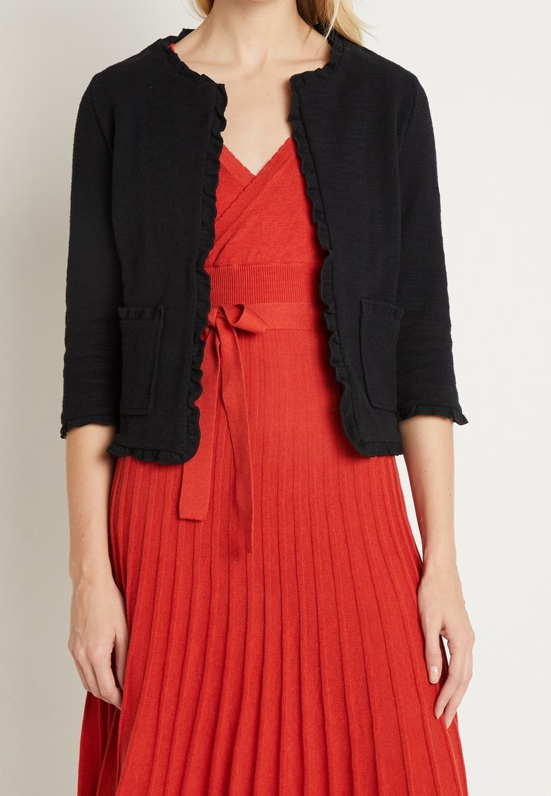 Femme portant un cardigan noir à volants sur une robe rouge plissée ceinturée, debout devant un fond clair uni.