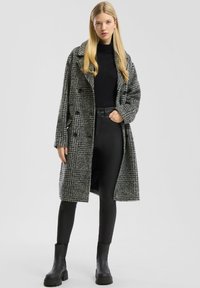 Manteau long à carreaux gris et noirs avec un design à double boutonnage, grands revers et tissu texturé ; associé à un haut noir et un pantalon.