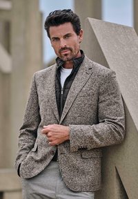 Braun karierter Hahnentritt-Blazer aus strukturiertem Stoff, darüber eine schwarze gesteppte Jacke getragen. Das Modell steht vor einer grauen geometrischen Struktur.