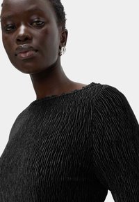 Czarny top o teksturowanej powierzchni z prążkowanym wzorem, z dekoltem w kształcie łodzi. Modelka nosi srebrne kolczyki w kształcie kół. Tło jest jednolicie białe.