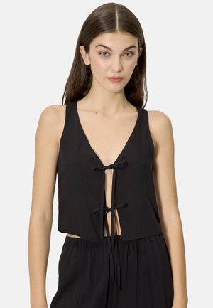 Mujer joven con cabello castaño lacio, vistiendo un top negro sin mangas y corto con cierres frontales de lazo, y pantalones negros de talle alto a juego.