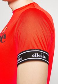 Rød træningsskjorte med en blank finish og et sort elastisk bånd med "ellesse" logo. Korte ærmer med rund halsudskæring.