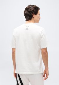 Άνδρας που κοιτάζει προς τα πίσω, φοράει λευκό μπλουζάκι με κοντά μανίκια, μικρό μαύρο λογότυπο και το κείμενο “ADP10” στο πάνω μέρος της πλάτης, συνδυασμένο με λευκό παντελόνι.